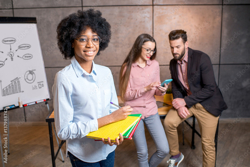 Portrait of a beautiful african businesswoman holding colorful books at the office with coworkers on