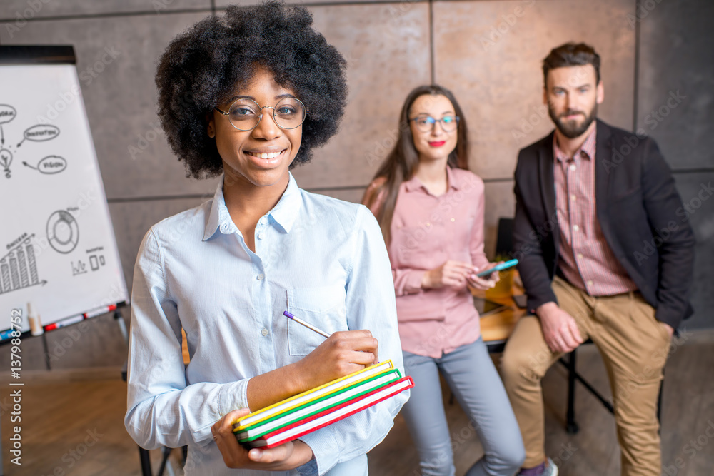 Portrait of a beautiful african businesswoman holding colorful books at the office with coworkers on