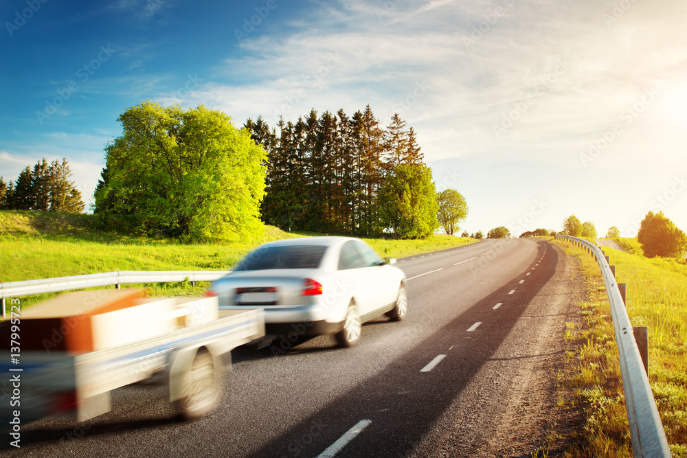 asphalt road on dandelion field with a car with trailer. vehicle moving on sunny evening