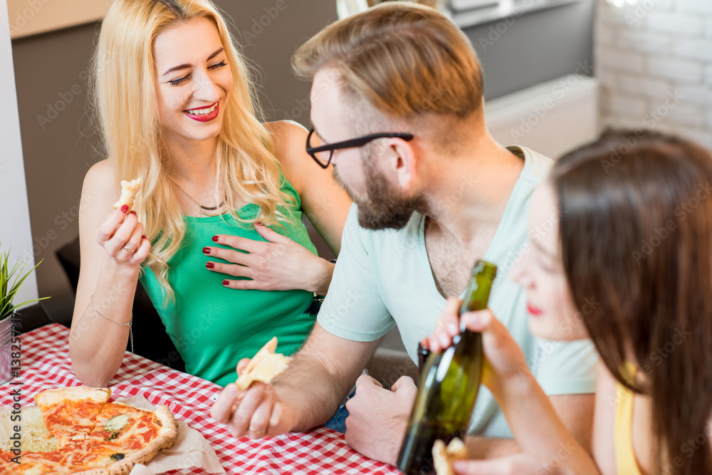 Young friends dressed casually in colorful t-shirts having lunch with pizza and beer at home