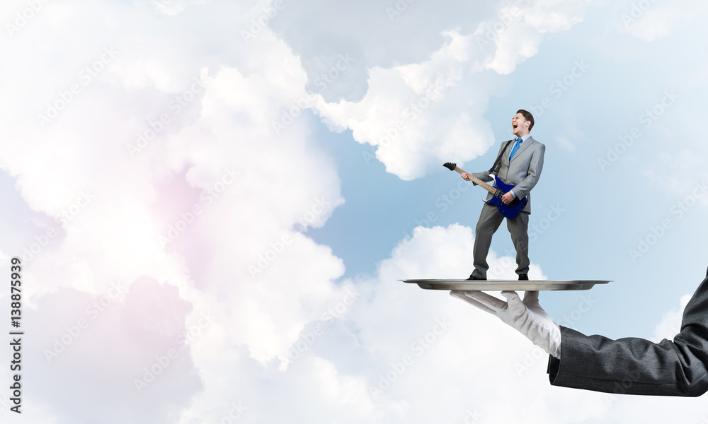 Businessman on metal tray playing electric guitar against blue sky background