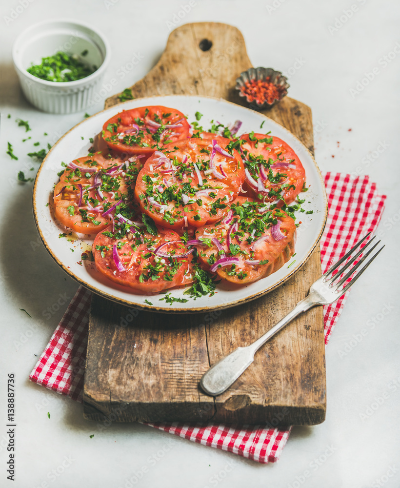 Fresh heirloom tomato, parsley and onion salad in white round plate on wooden board over light grey 