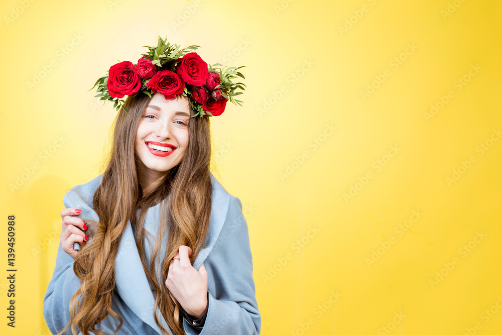 Colorful portrait of a beautiful woman in blue coat with wreath made of red roses on the yellow back