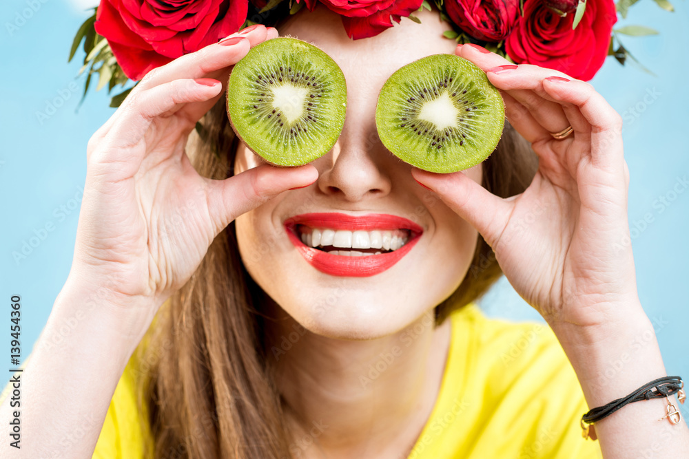 Colorful portrait of a beautiful woman with flower wreath holding slices of kiwi fruit on the blue b