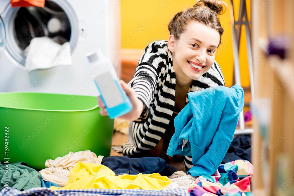 Young smiling housewife holding a bottle with detergent and t-shirt sitting on the floor near the wa