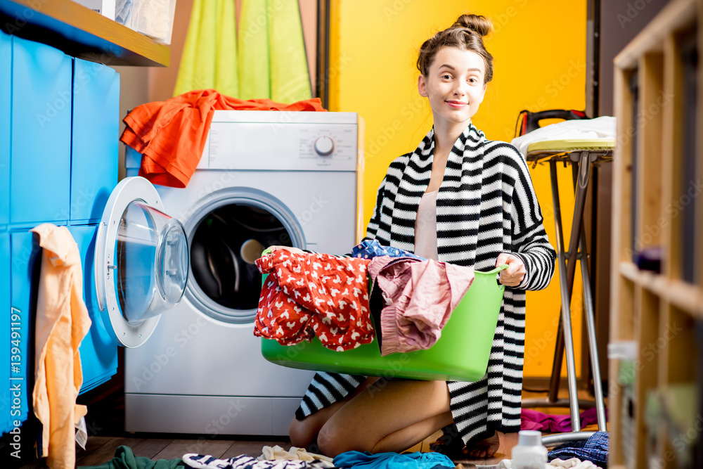 Young housewife holding a basket with clothes near the washing machine sitting on the floor at home