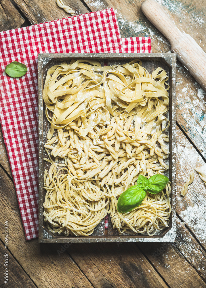 Various homemade fresh uncooked Italian pasta with flour and green basil leaves in wooden tray over 