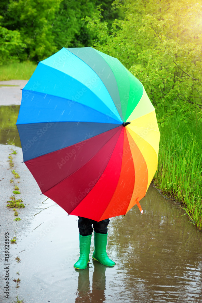 Child walking in wellies in puddle on rainy weather. Boy holding colourful umbrella under rain in su