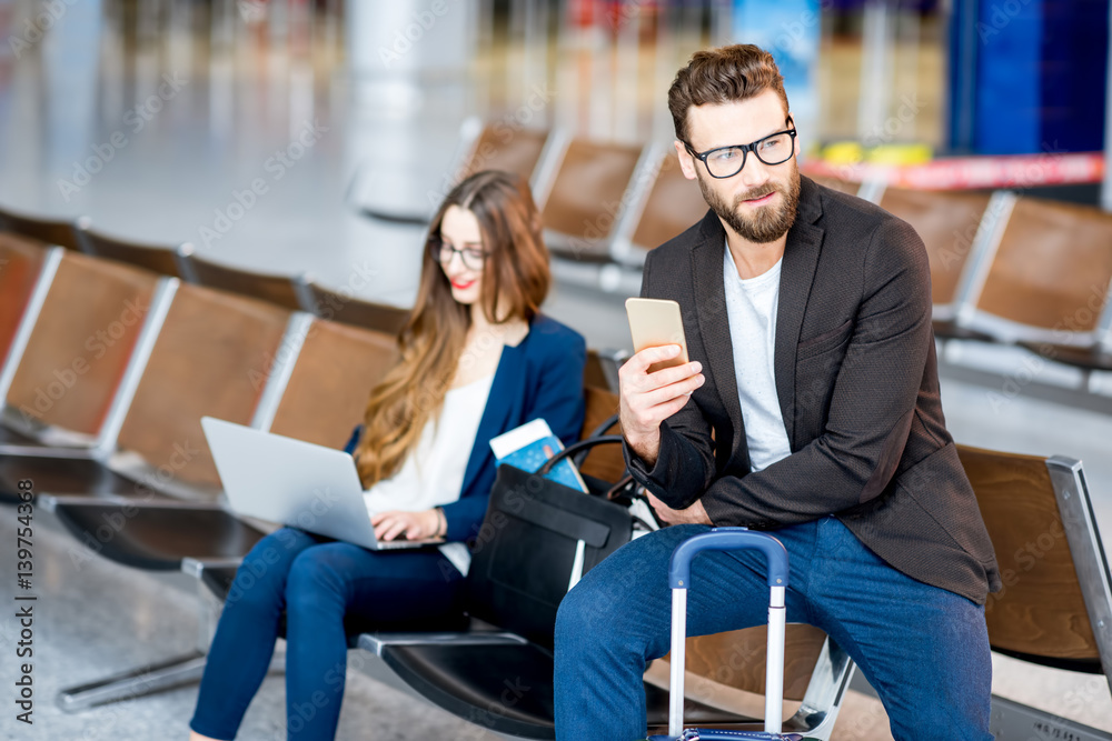 Elegant business couple sitting with laptop, phone and suitcase at the waiting hall in the airport. 