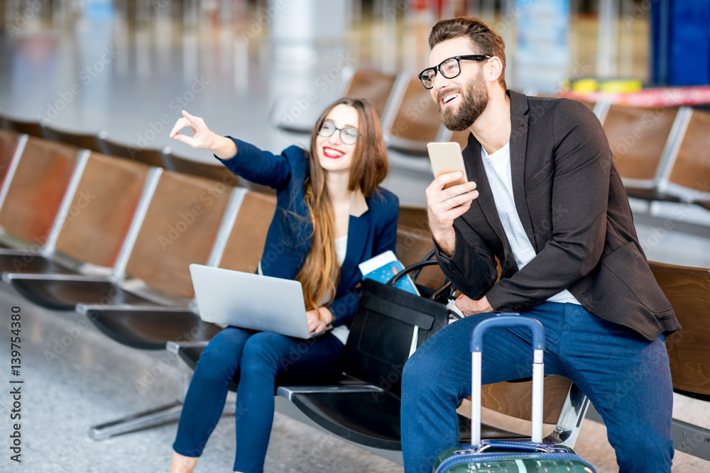 Elegant business couple sitting with laptop, phone and suitcase at the waiting hall in the airport. 