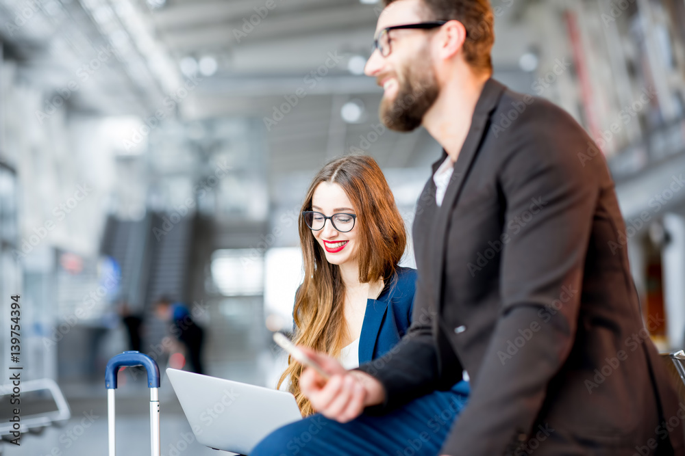 Elegant business couple sitting with laptop, phone and suitcase at the waiting hall in the airport. 