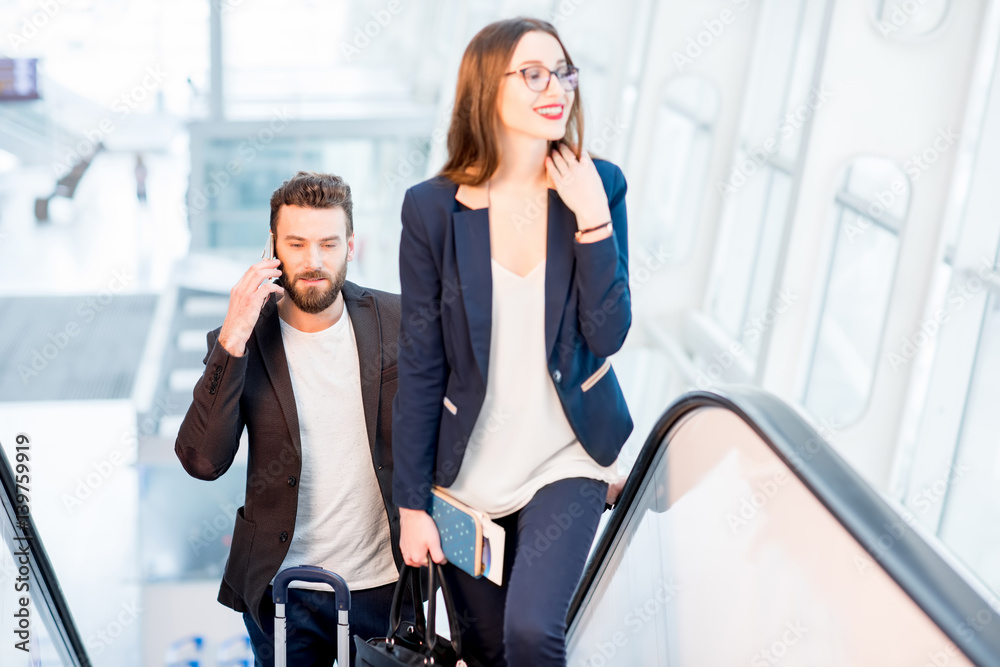 Business couple talking with phones while getting up on the escalators with baggage at the airport