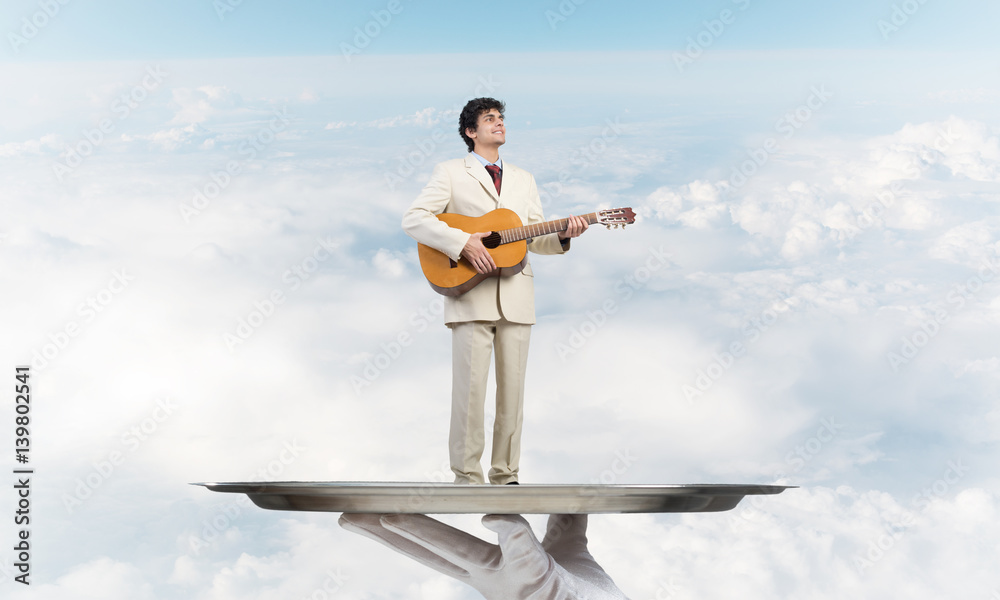 Businessman on metal tray playing acoustic guitar against blue sky background