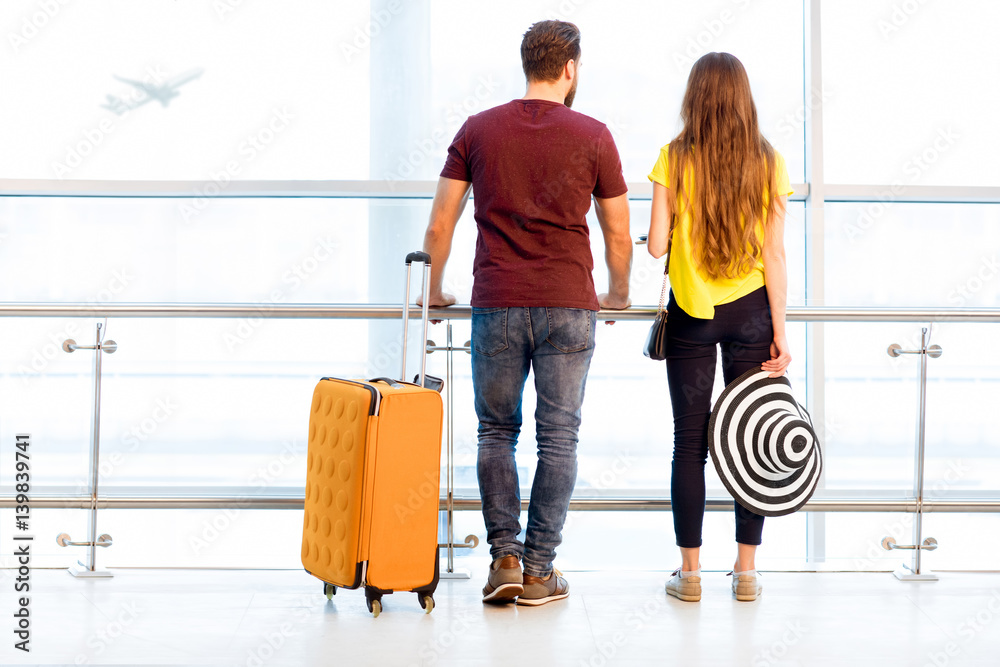 Young couple in colorful t-shirts waiting with baggage near the window at the departure area of the 