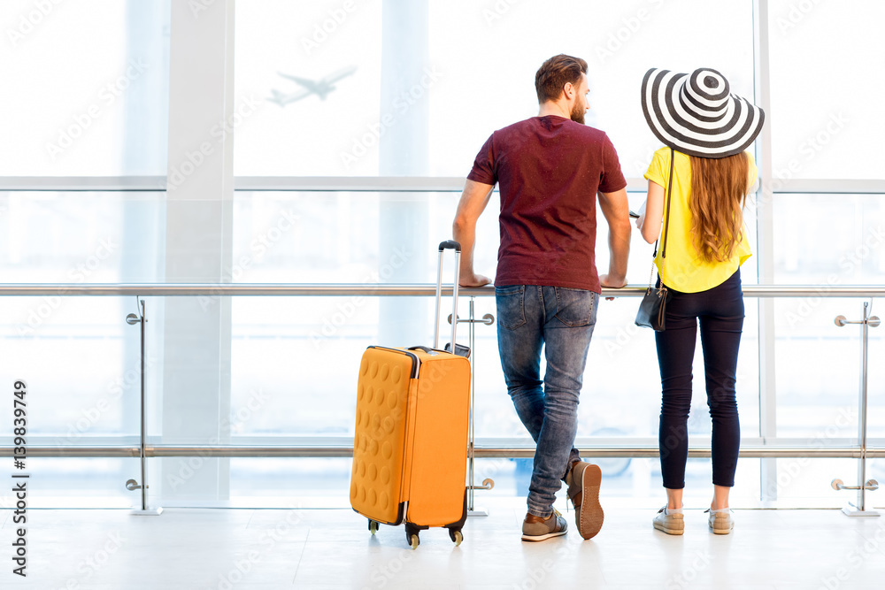 Young couple in colorful t-shirts waiting with baggage near the window at the departure area of the 