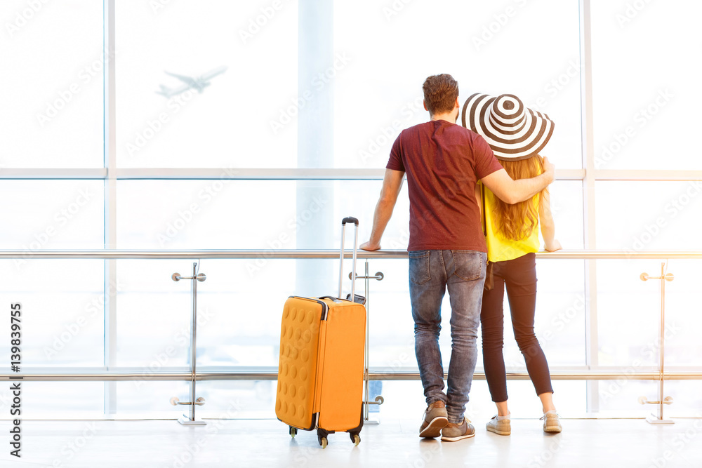 Young couple in colorful t-shirts waiting with baggage near the window at the departure area of the 