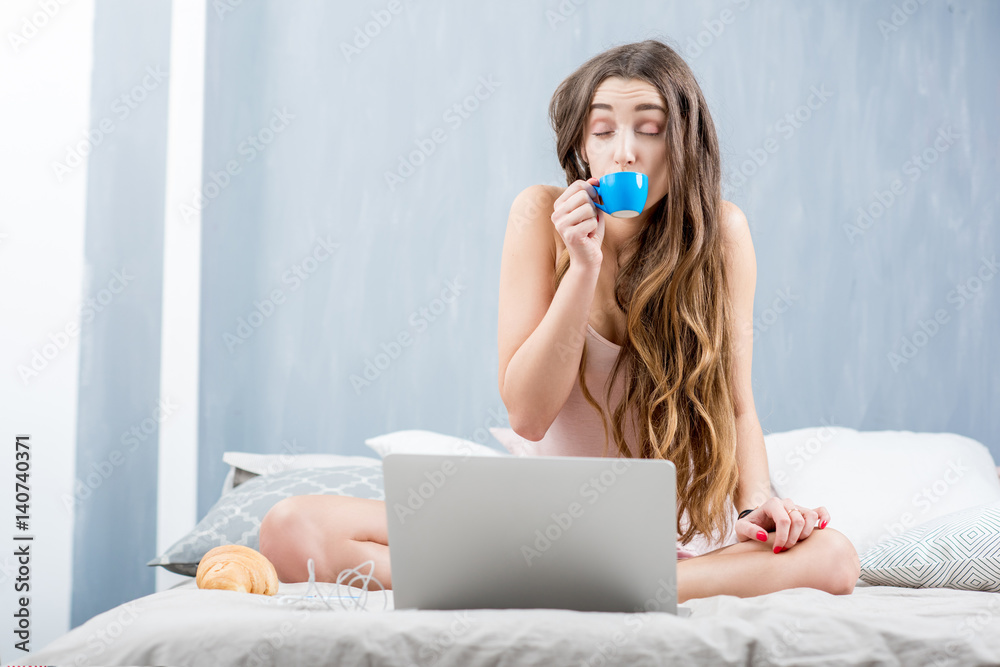 Young woman in sleepwear drinking coffee sitting with laptop in the bed