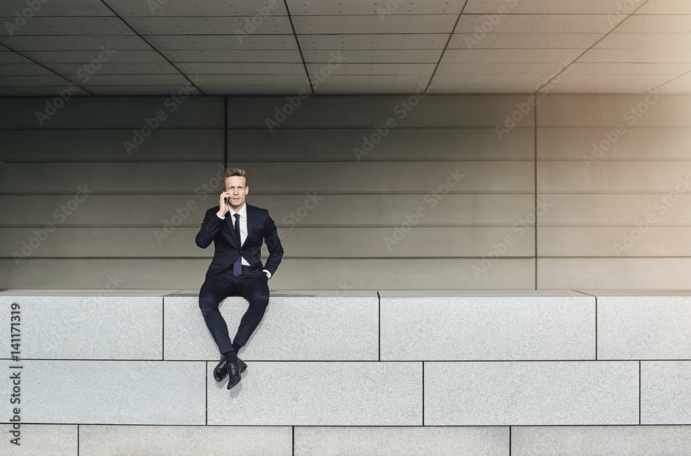 Businessman speaking phone stitting on brick wall