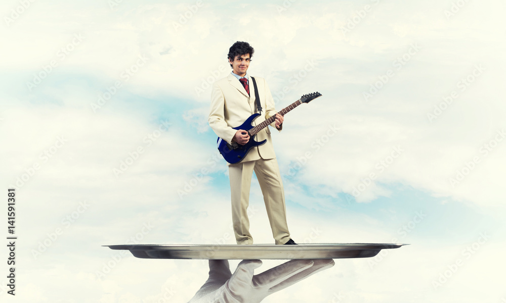 Businessman on metal tray playing electric guitar against blue sky background