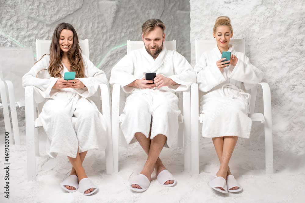 Three friends in bathrobes sitting with phones, applying salt therapy in the salt room