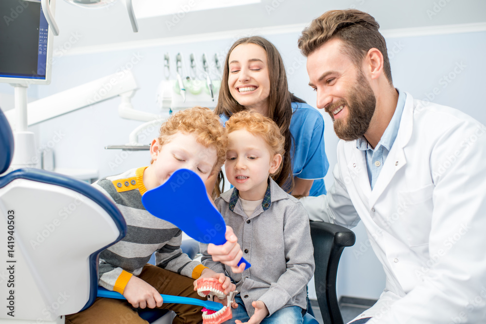 Young boys looking at the mirror with toothy smile sitting with dentist and woman assistant at the d