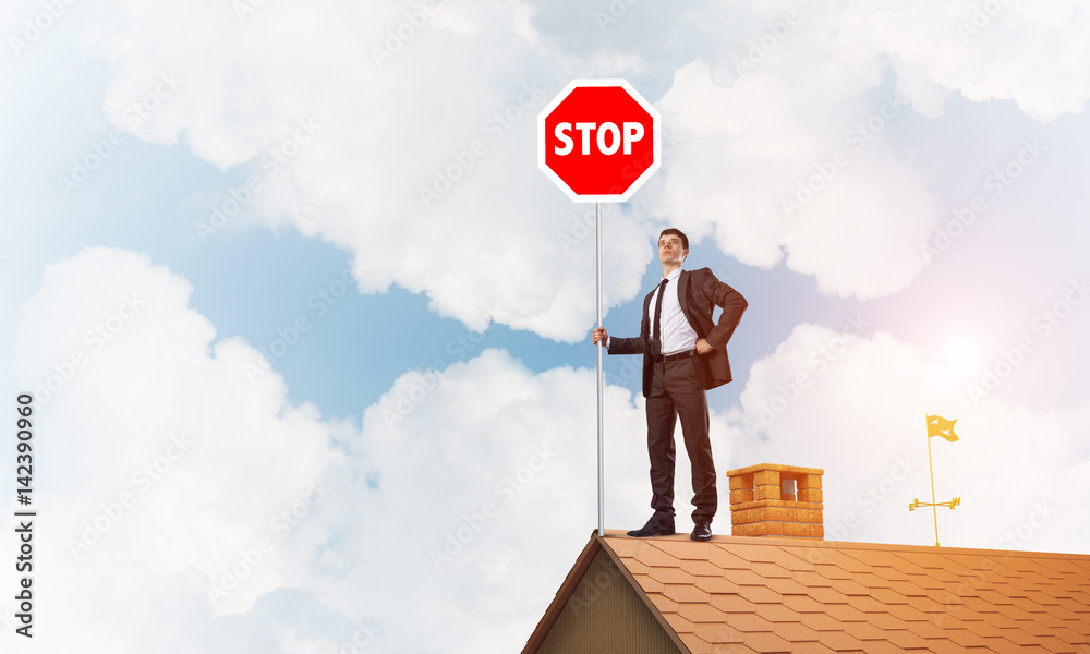 Caucasian businessman on brick house roof showing stop road sign. Mixed media