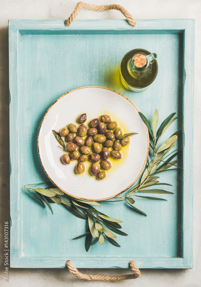 Pickled green Mediterranean olives on white ceramic plate, olive tree branch and virgin olive oil in