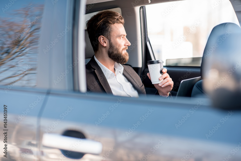 Handsome businessman sitting with coffee to go on the backseat of the car