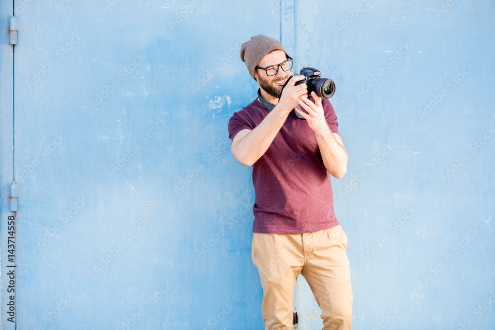 Portrait of a stylish photographer dressed casual in t-shirt and hat standing with camera on the blu