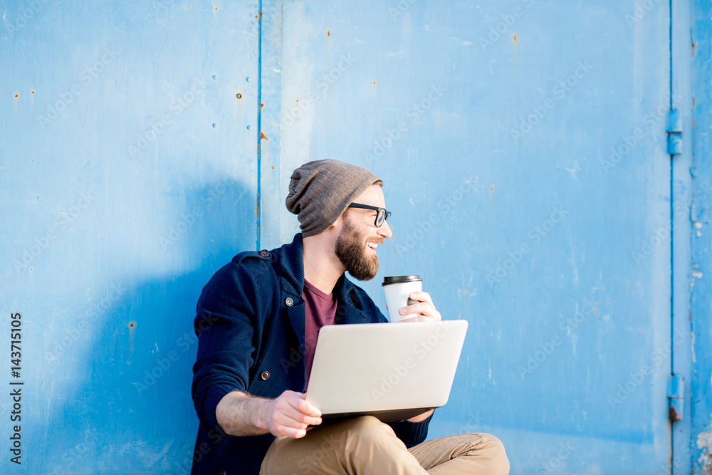 Stylish man dressed casual in sweater and hat working with laptop sitting with coffee near the blue 