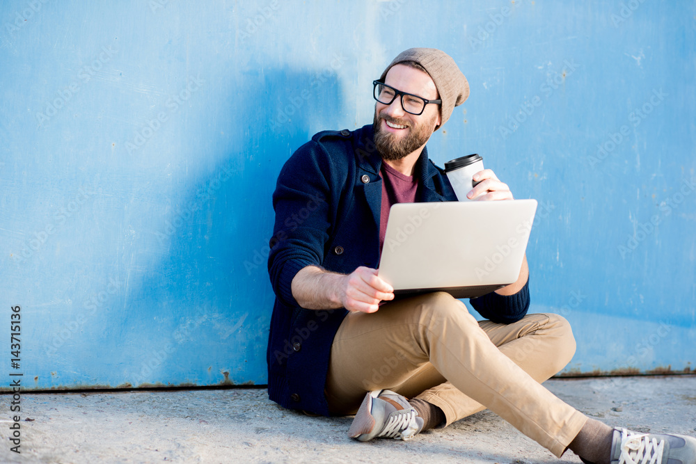 Stylish man dressed casual in sweater and hat working with laptop sitting with coffee near the blue 