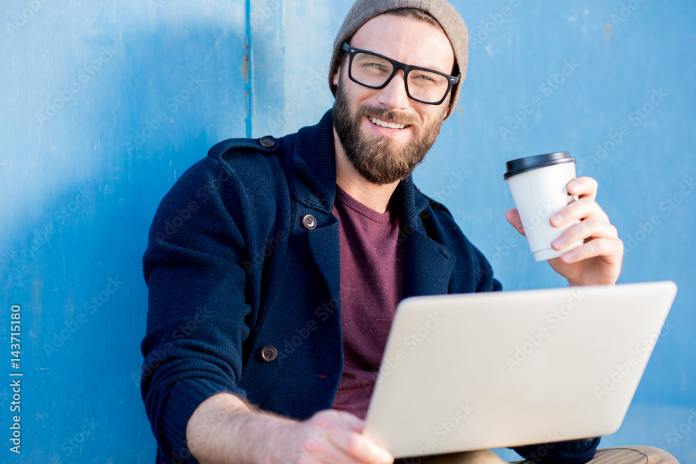 Stylish man dressed casual in sweater and hat working with laptop sitting with coffee near the blue 