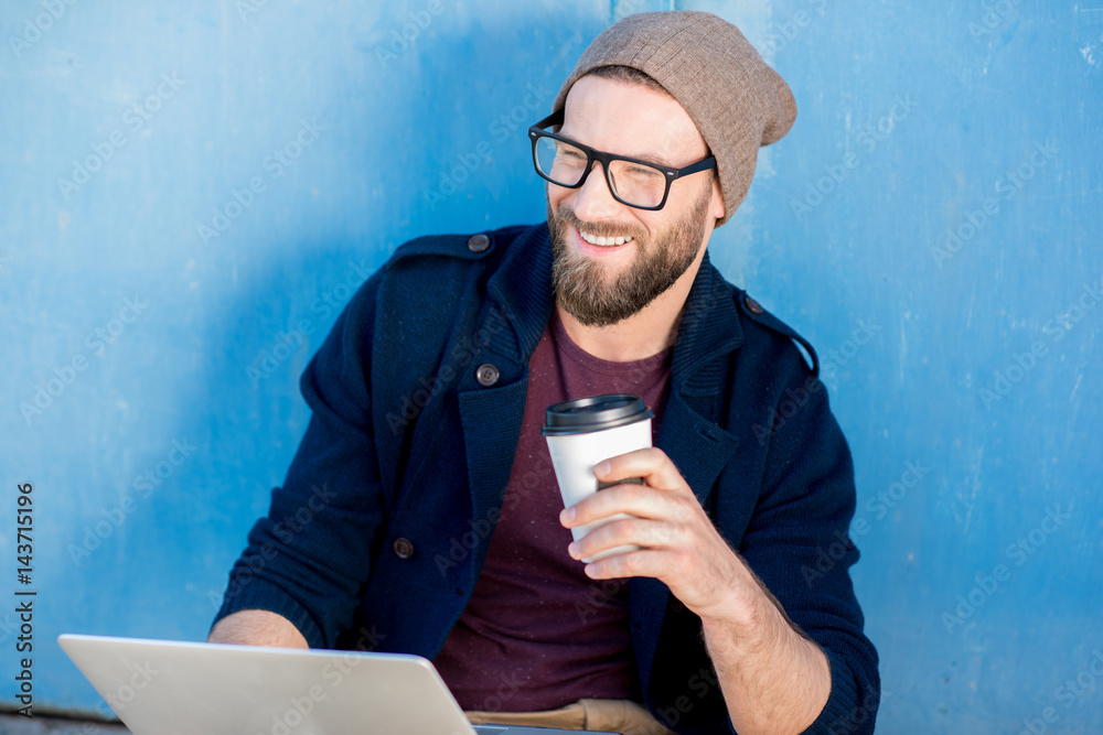 Stylish man dressed casual in sweater and hat working with laptop sitting with coffee near the blue 