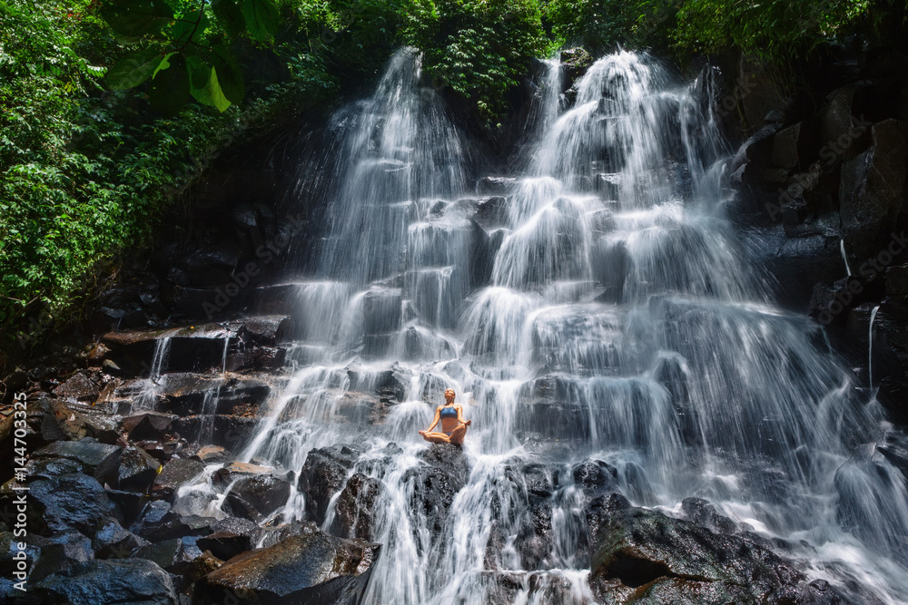 Beautiful happy woman travel in Bali jungle. Sit in zen-like yoga pose under flowing spring water, e