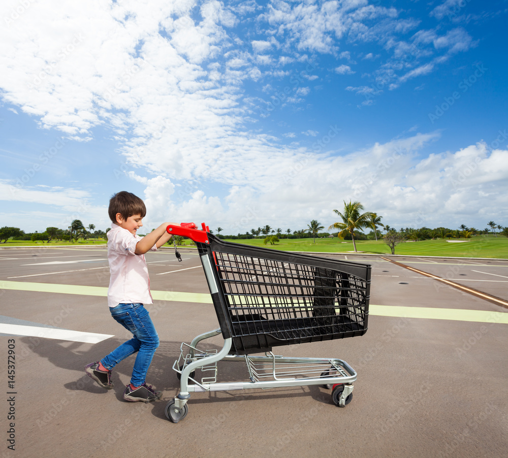 Kid boy pushing empty shopping cart at parking lot