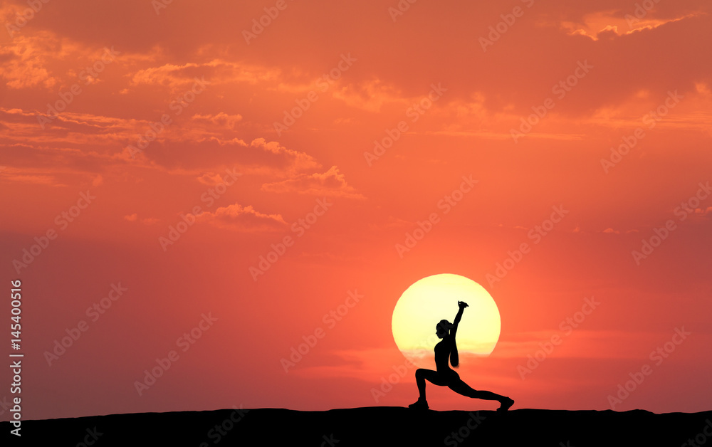 Silhouette of a standing sporty woman practicing yoga with raised up arms on the hill on the backgro