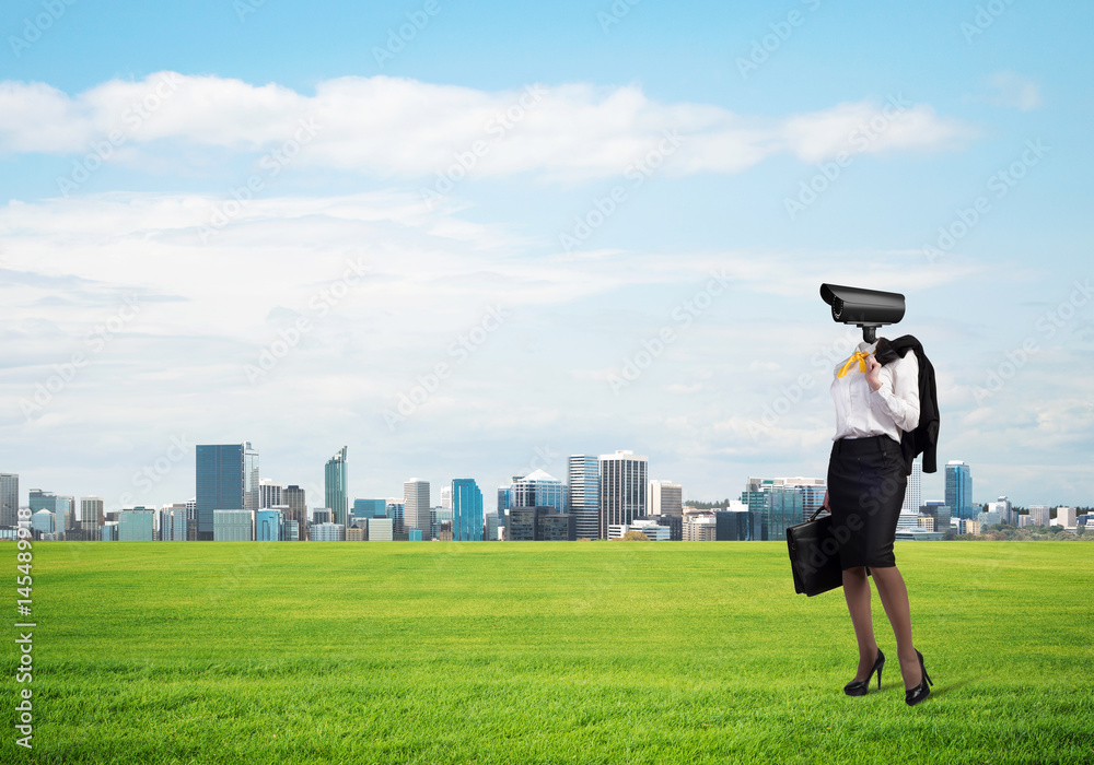 Camera headed woman standing on green grass against modern cityscape