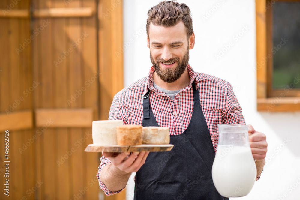 Portrait of a handsome farmer in apron standing with goat cheeses and milk outdoors on the rural hou