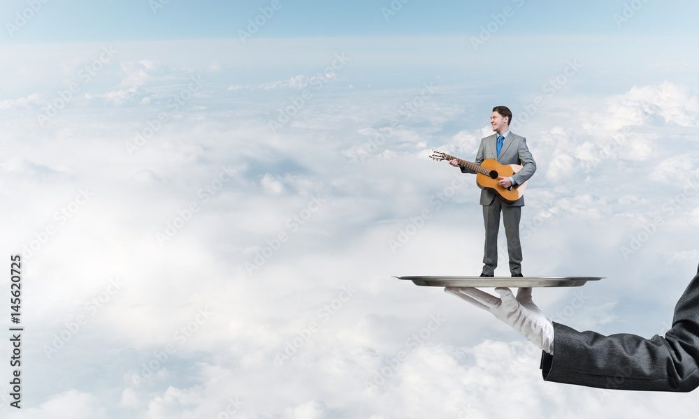 Businessman on metal tray playing acoustic guitar against blue sky background