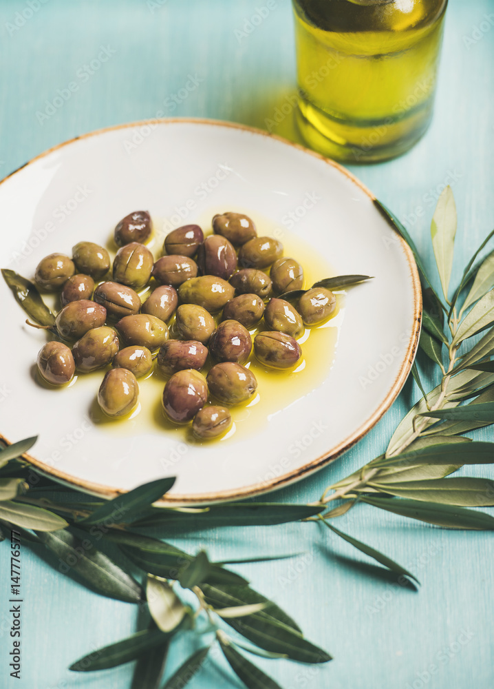 Pickled green Mediterranean olives on white ceramic plate, olive tree branch and virgin olive oil in