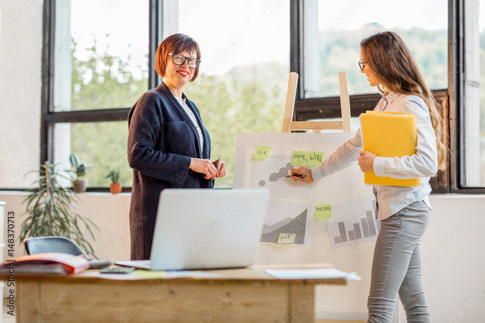 Older and young businesswomen having a conversation near the board with financial charts at the offi