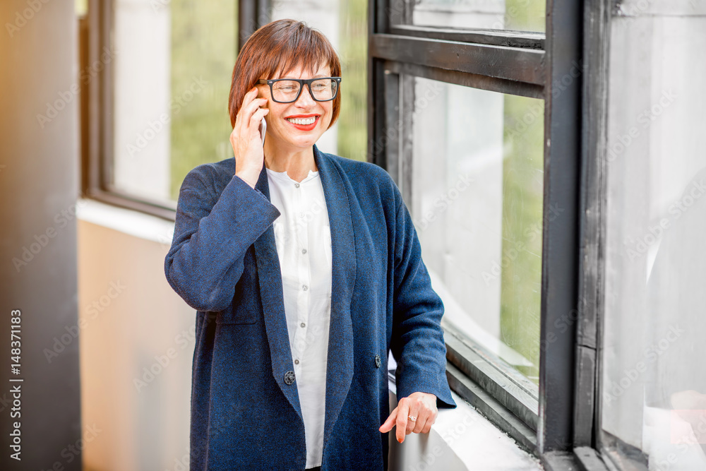 年长优雅的女商人站在室内靠窗打电话