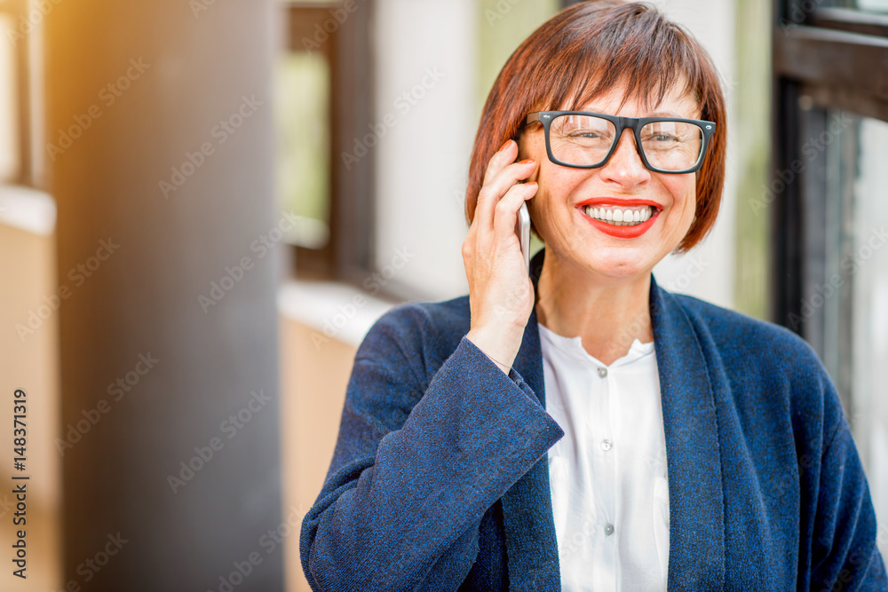 Older and elegant businesswoman talking phone standing indoors near the window