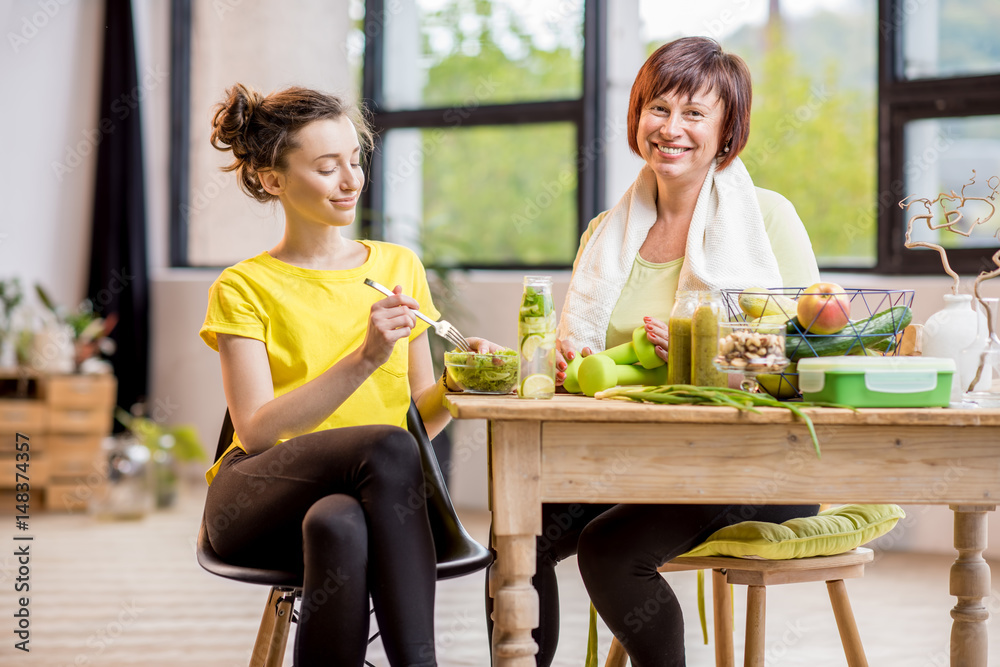 Young and older women exercising with dumbbells and eating healthy food indoors on the window backgr