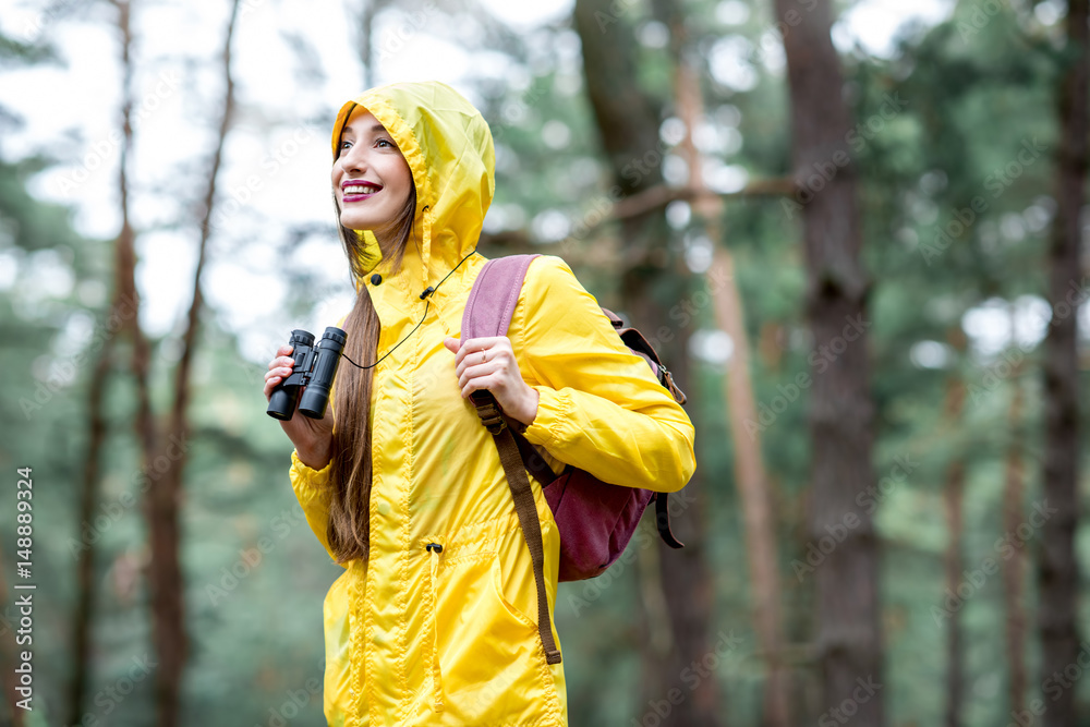 Young woman in yellow raincoat hiking with binoculars and backpack in the green pine forest. Bird wa