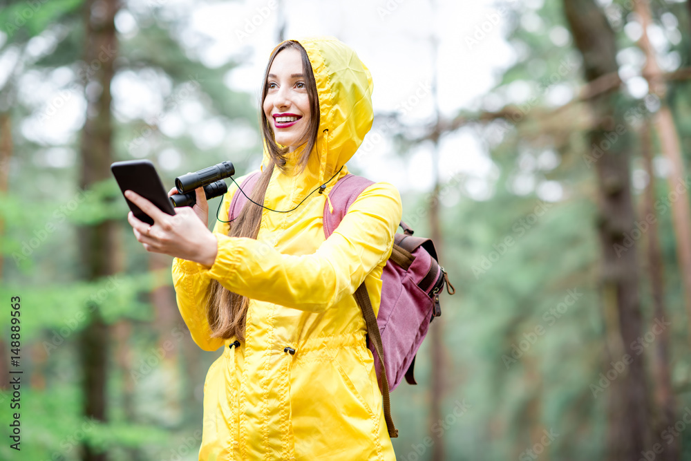 穿着黄色雨衣的年轻女子在绿色松树f中行走时使用智能手机进行定向