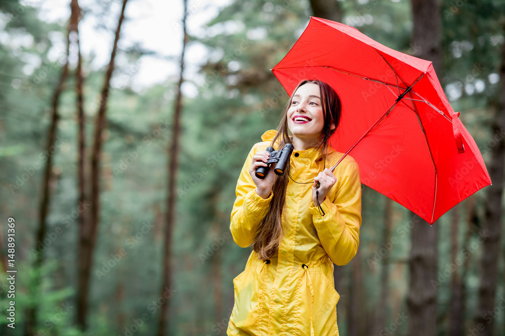 身穿黄色雨衣的年轻女子带着双筒望远镜和红色雨伞在松林中徒步旅行