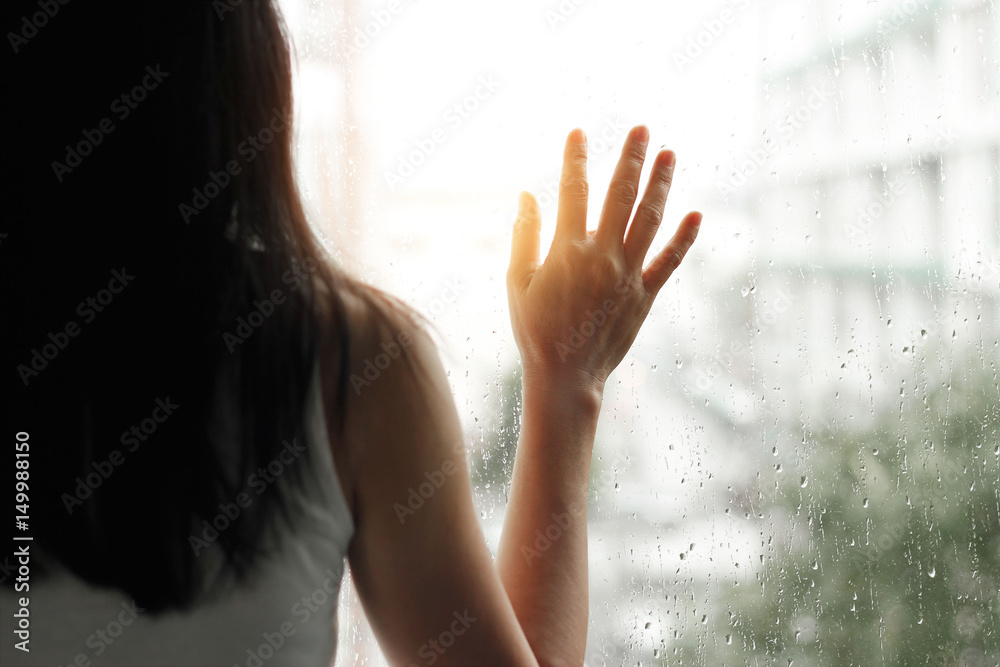 悲伤的女人透过玻璃窗看着城市背景上的雨滴