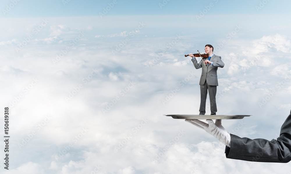 Businessman on metal tray playing violin against blue sky background