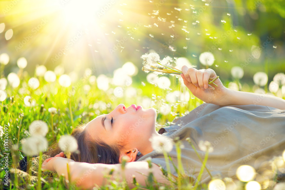 Beautiful young woman lying on the field in green grass and blowing dandelion